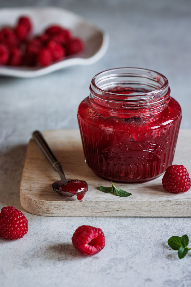 Samtige Himbeerkonfitüre ohne Kerne in einem Glas mit einem Löffel auf einem Holzbrett mit Himbeeren im Vorder- und Hintergrund.