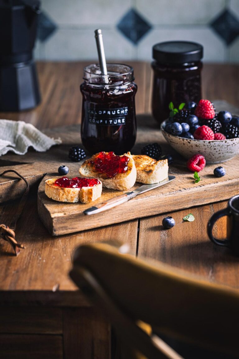 Waldfrucht Marmelade ohne Kerne auf gerösteten Baguettescheiben auf einem Holzbrett mit Beeren und Marmeladengläsern im Hintergrund.