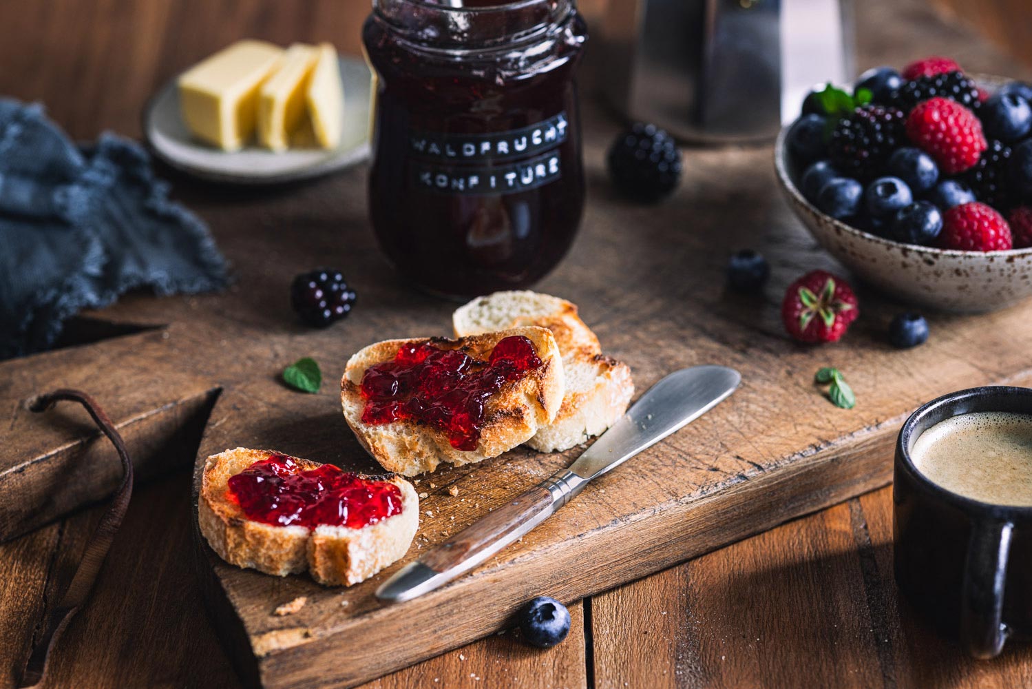 Waldfrucht Marmelade ohne Kerne auf gerösteten Baguettescheiben auf einem Holzbrett mit Beeren und Marmeladengläsern im Hintergrund.