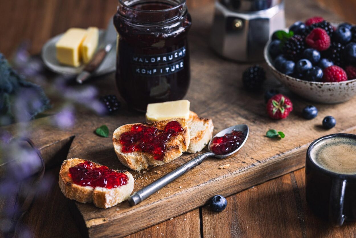 Waldfrucht Marmelade ohne Kerne auf gerösteten Baguettescheiben auf einem Holzbrett mit Beeren und Marmeladengläsern im Hintergrund.