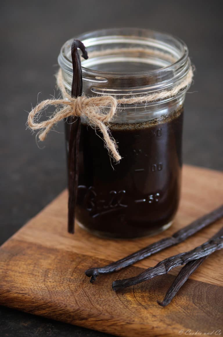 Vanilleextrakt in einem Glas auf einem Holzbrett mit Vanilleschoten im Vordergrund.