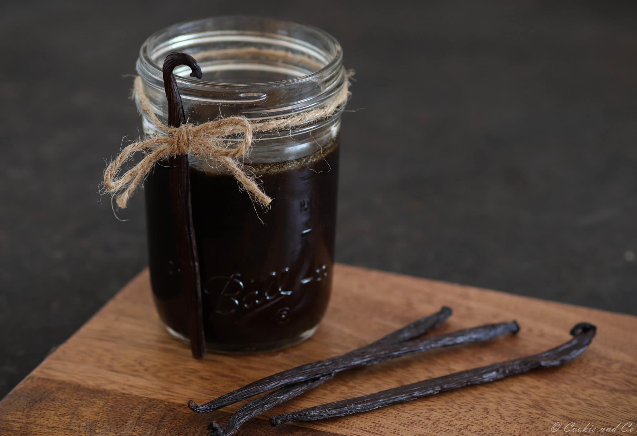 Vanilleextrakt in einem Glas auf einem Holzbrett mit Vanilleschoten im Vordergrund.