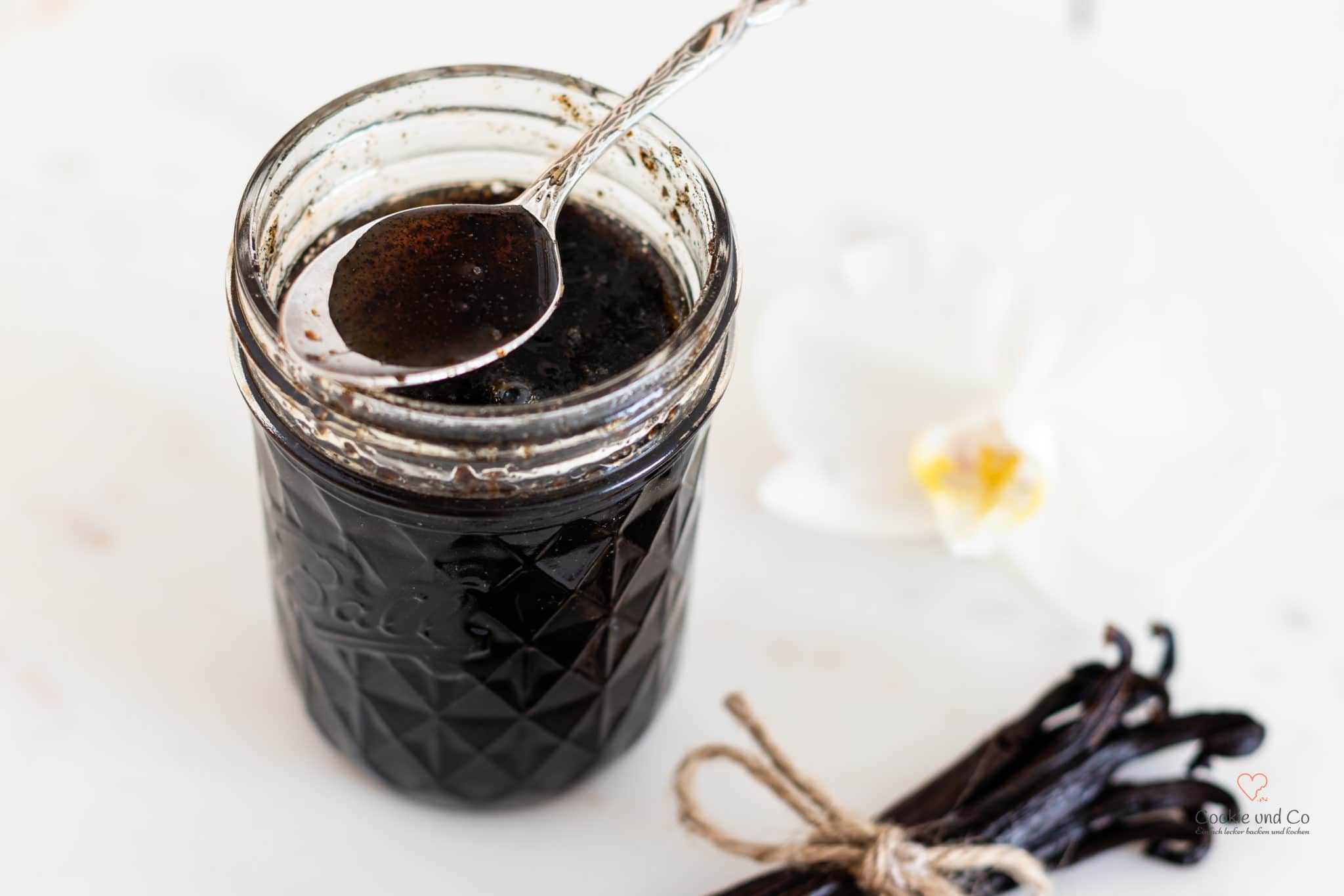 Selbst gemachter Vanilleextrakt in einem Einmachglas mit Vanilleschoten und einer Orchideenblüte daneben.