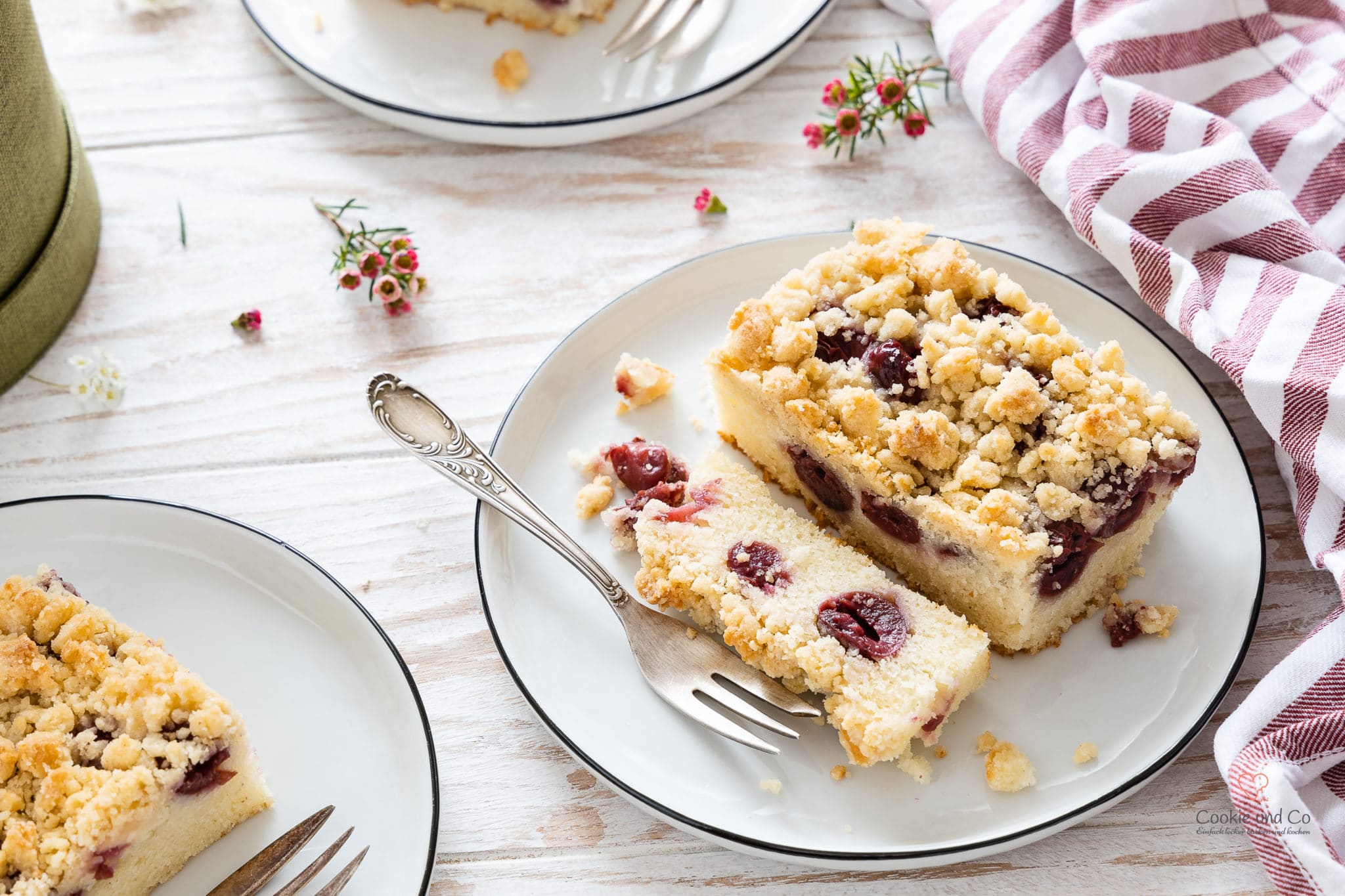 Ein Kirsch-Streuselkuchen auf einem Teller mit einer Gabel und Blumen im Hintergrund (schneller Blechkuchen aus Rührteig)