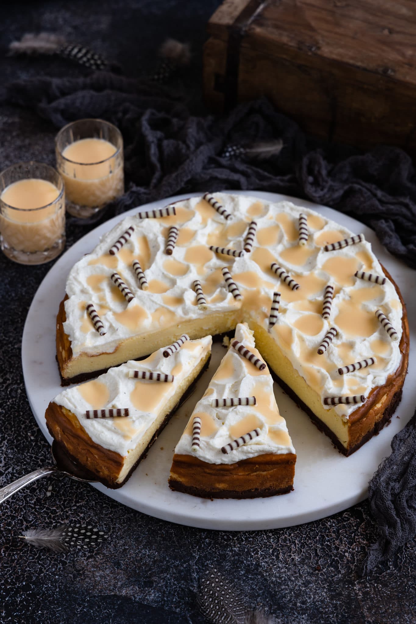 Eierlikör Käsekuchen auf einem Marmorteller mit Eierlikörgläsern und einer Holzkiste im Hintergrund.