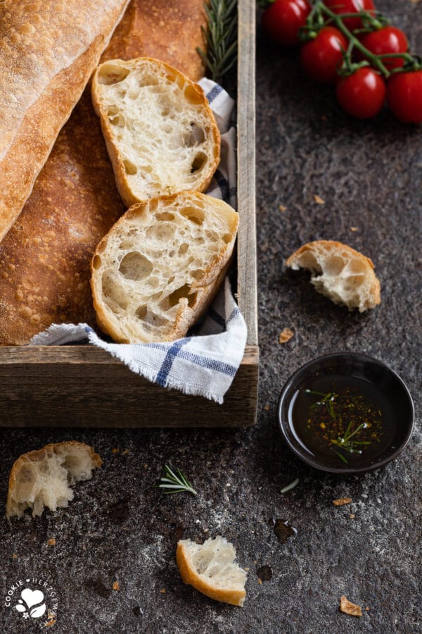 Ciabatta aufgeschnitten in einer Holzkiste mit Olivenöl und Tomaten im Hintergrund.