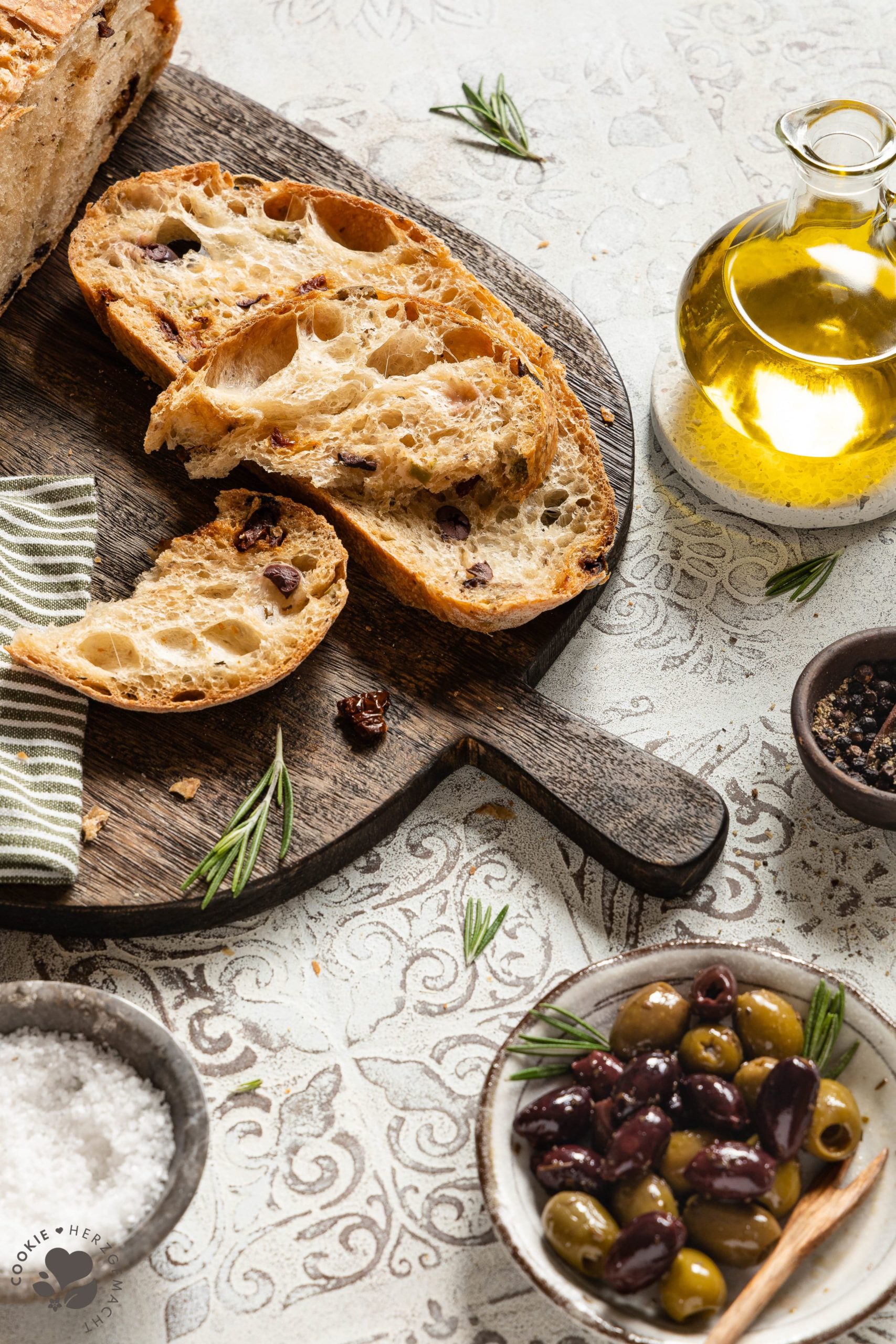 Mediterranes Olivenbrot aufgeschnitten auf einem Holzbrett mit einer Schale Oliven und einer Flasche Olivenöl.