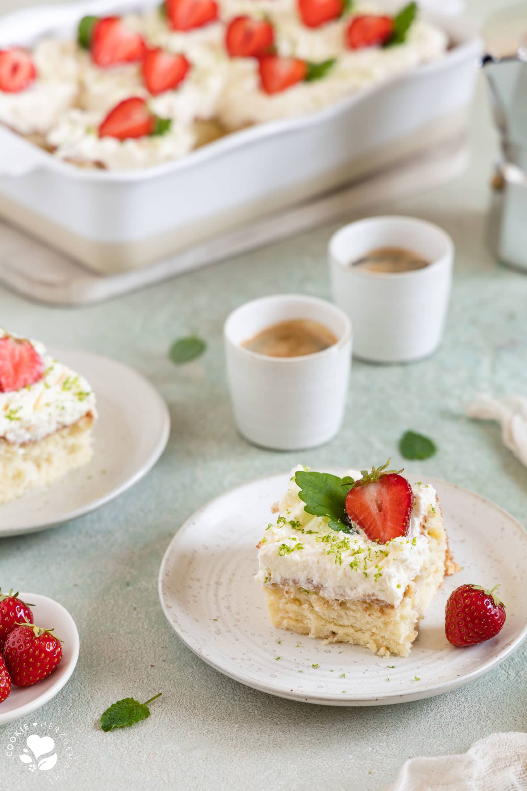 Ein Stück mexikanischer Tres leche Kuchen (Milchkuchen) mit Erdbeeren auf einem Teller mit Espressotassen und einer Backform mit Kuchen im Hintergrund.