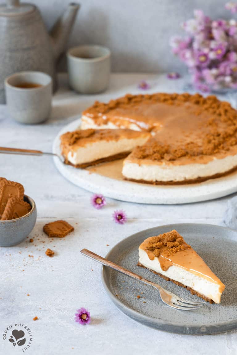 Ein Stück Karamell-Käsekuchen ohne Backen auf einem Teller mit einer Kuchengabel und dem Kuchen und Kaffeetassen im Hintergrund.