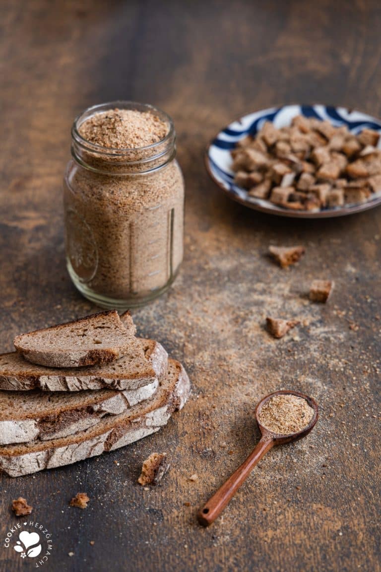 Gemahlenes Altbrot in einem Glas mit einem Löffel im Vordergrund und getrockneten Brotwürfeln im Hintergrund.