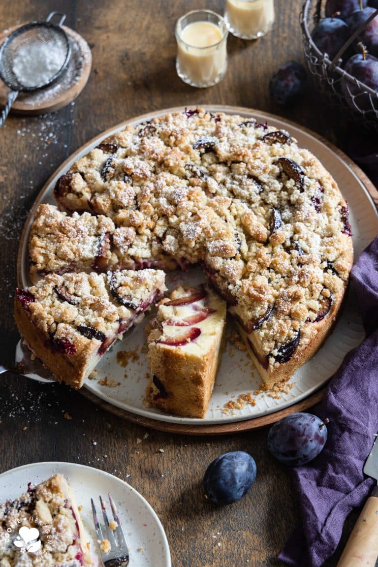 Ein Zwetschgenkuchen mit Streuseln auf einer Tortenplatte mit einem Puderzuckersieb, zwei Gläsern mit Eierlikör im Hintergrund und einem Korb mit Pflaumen.