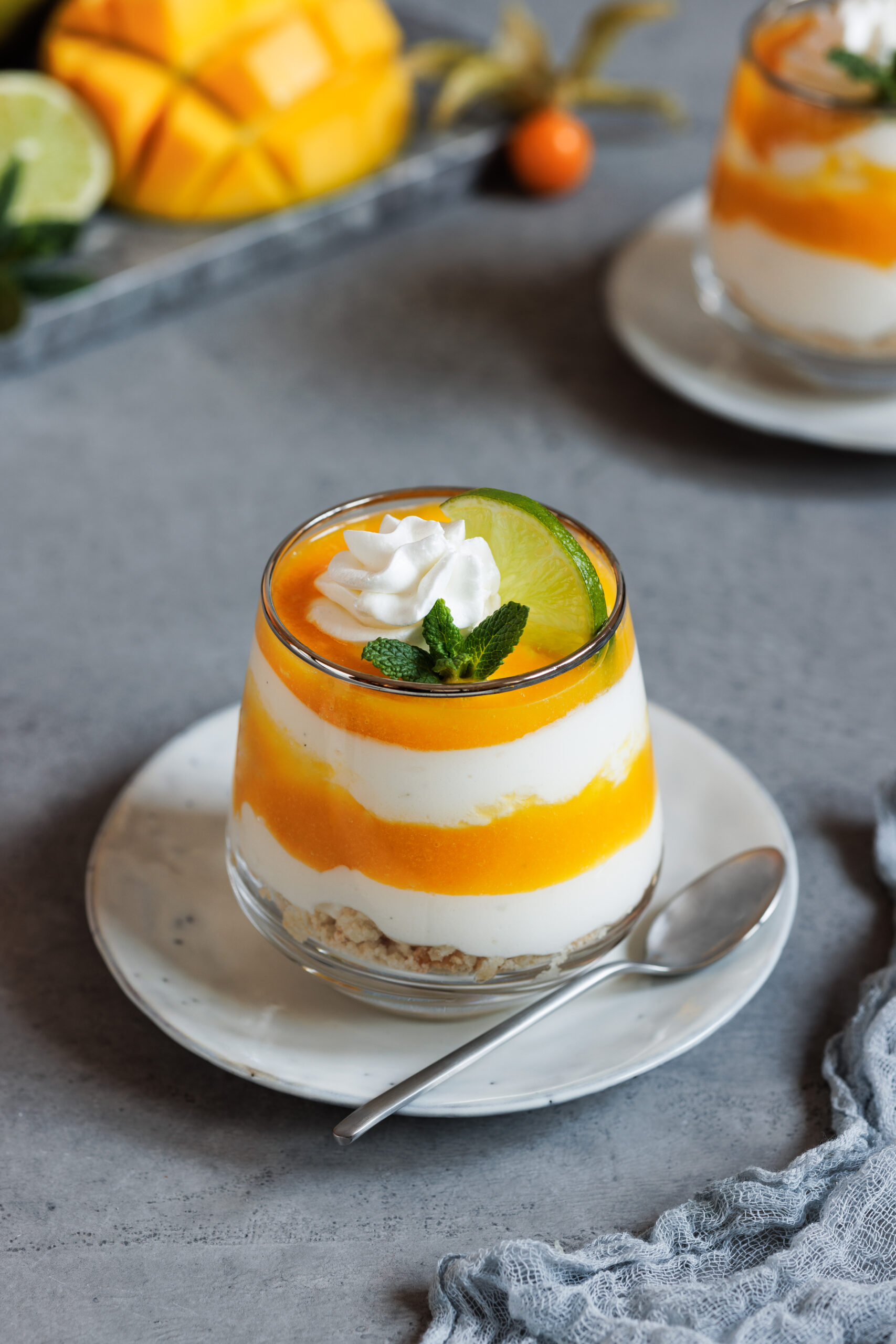 Schichtdessert mit Mango und Jog´hurt in einem Glas auf einem Teller mit einem Löffel.