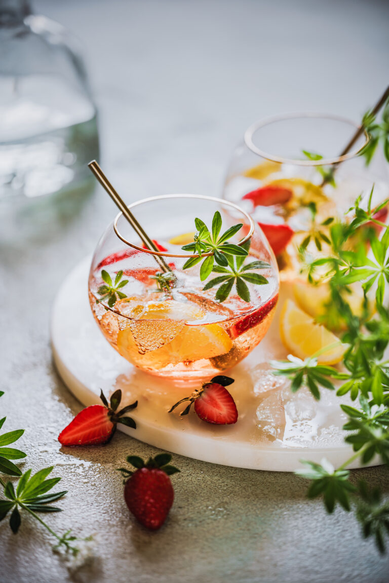 Maibowle mit Erdbeeren, Zitrone und Waldmeister in zwei Bowlegläsern auf einem Marmortablett mit Waldmeister und Erdbeeren im Vordergrund.