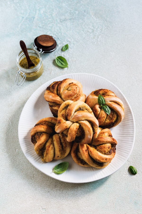 Mit Pesto gefüllte Brötchen-Knoten auf einem Teller mit einem Glas grünem Pesto und Basilikumblättern im Hintergrund.