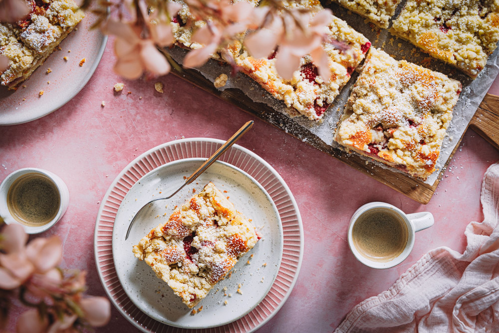 Ein Stück Rhabarber-Erdbeer-Kuchen mit Streuseln auf einem Teller mit zwei Tassen Kaffee daneben und einem Brett mit weiteren Kuchenstücken im Hintergrund.