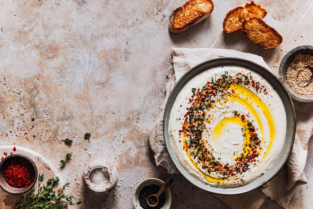 Feta-Dip in einer Schüssel mit Toppings sowie Schälchen mit Gewürzen und geröstetem Brot daneben.