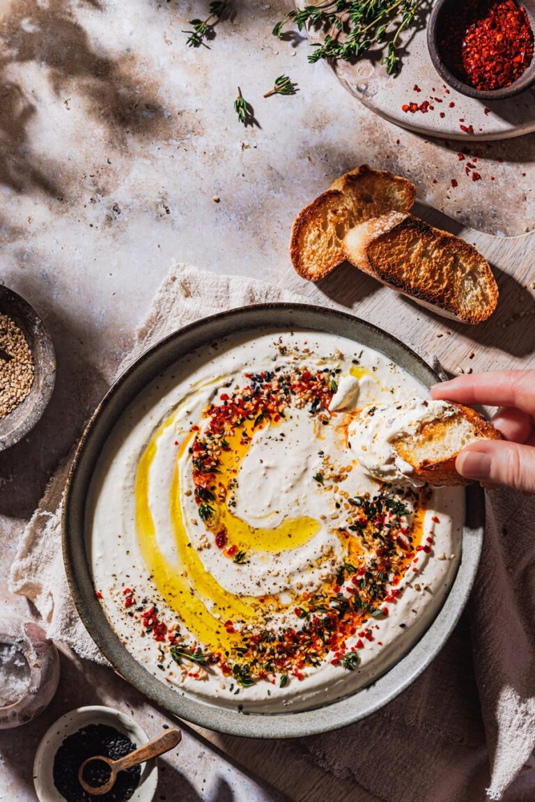 Feta-Dip in einer Schüssel mit Toppings, in die eine Hand Brot hineindipt sowie Schälchen mit Gewürzen und geröstetem Brot daneben.