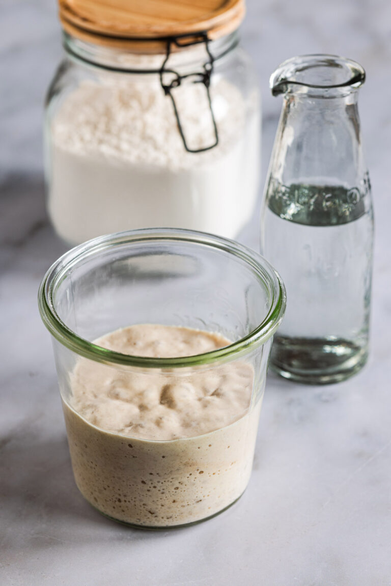 Poolish, ein Hefevorteig i einem Einmachglas nit einer Karaffe Wasser und einem Glas Mehl im Hintegrund.