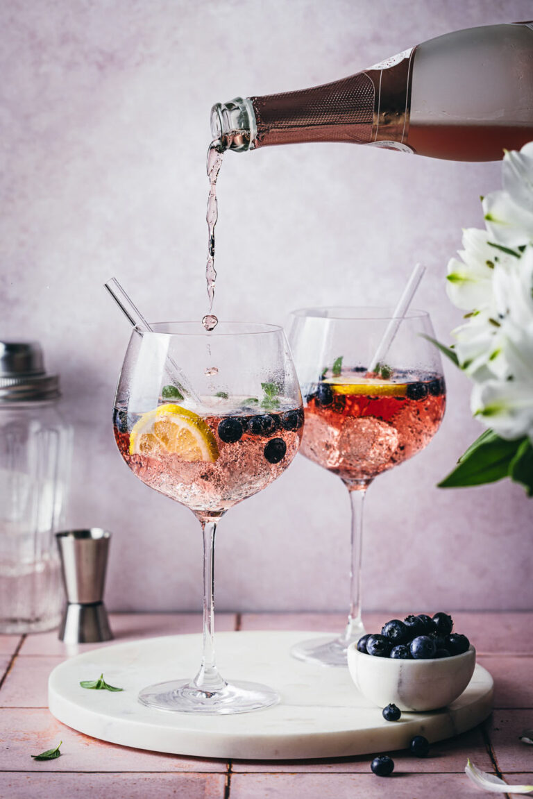 Blueberry Gin Spritz in zwei Cocktailgläsern auf einer Marmorplatte mit weißen Blumen und frischen Heidelbeeren im Vordergrund. Im Hintergrund stehen ein Jigger und ein Cocktailshaker.