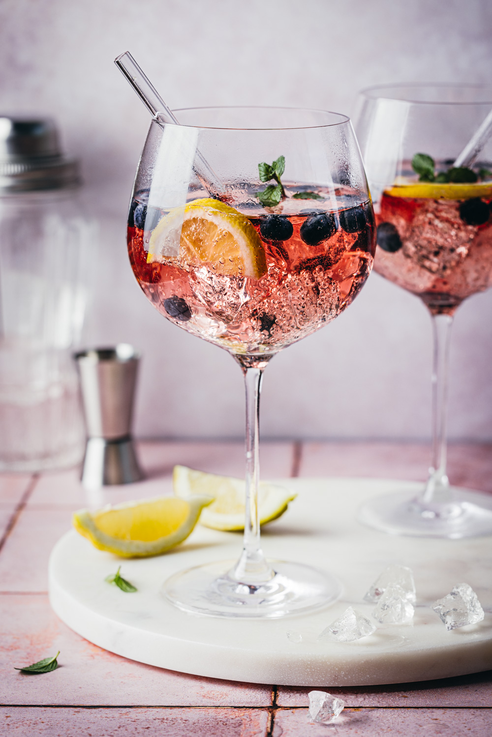 Blueberry Gin Spritz in zwei Cocktailgläsern auf einer Marmorplatte mit wZitronenspalten und Eiswürfeln im Vordergrund. Im Hintergrund stehen ein Jigger und ein Cocktailshaker.