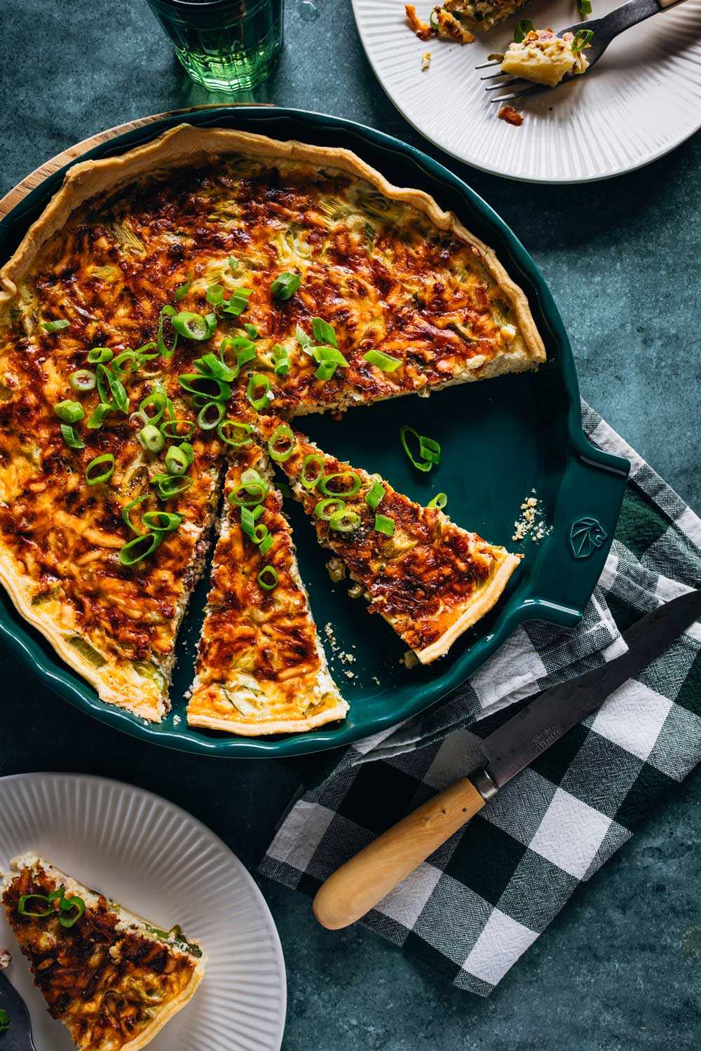 Eine frisch gebackene Quiche Lorraine in einer grünen Tarteform, aufgeschnitten und garniert mit geschnittenen Frühlingszwiebeln. Einige Stücke sind entnommen und auf weißen Tellern serviert. Daneben liegt ein Messer auf einer grün-weiß karierten Serviette.